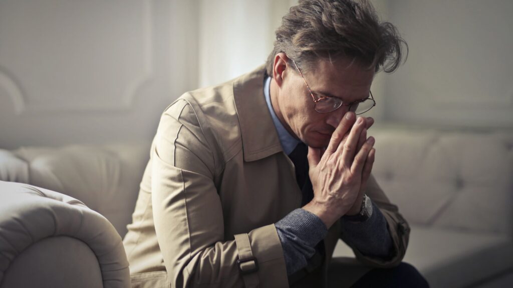 Fatigued man sitting on couch with hands on face.
