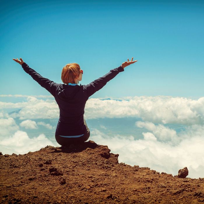 Woman sitting on a mountain peak with arms raised.